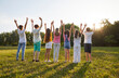 © Studio Romantic - Group of kids friends raising hands up on green grass in the park standing back in a line at sunset. Children having fun together outdoors on a sunny summer day in casual clothes in nature.