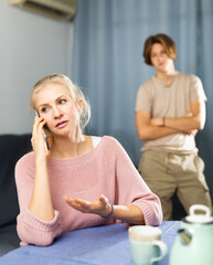 Wall Mural - Portrait of focused woman sitting in dining room and speaking on phone while her resentful or worried teenage son standing behind