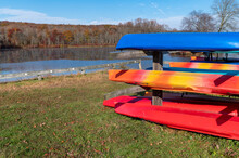 Racks Of Colorful Kayaks And Canoes By State Park Autumn Lake With Woodland Background And Copy Space