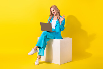 Poster - Full length photo of pleasant woman wear blue stylish suit sit on white cube talk on laptop video call isolated on yellow background