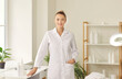 © Studio Romantic - Portrait of professional beautician at work. Happy female cosmetologist in her workplace. Beautiful young woman wearing white coat standing by table with beauty products, looking at camera and smiling