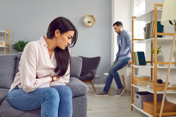 Wall Mural - Upset young woman sitting on the sofa and a man in background ignoring each other at home. Stressed married couple sitting separately in the living room. Quarrel and divorce concept.
