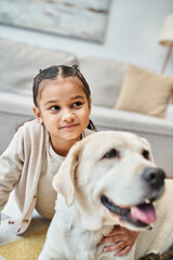 Wall Mural - happy cute girl sitting on carpet and stroking dog in modern living room, kid and labrador