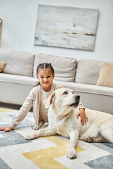 Wall Mural - happy cute girl sitting on carpet and stroking labrador in modern living room, kid and furry friend