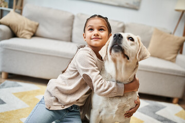 Wall Mural - happy cute elementary age girl looking up and hugging labrador in modern apartment, pet and child