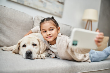 Wall Mural - cute elementary age girl taking selfie with labrador dog in modern apartment, domestic animal