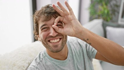 Wall Mural - Joyful young man sitting on cozy sofa with a cheerful smile, confidently making an ok sign with hand over his eye, playfully looking through fingers in the comfort of his home.