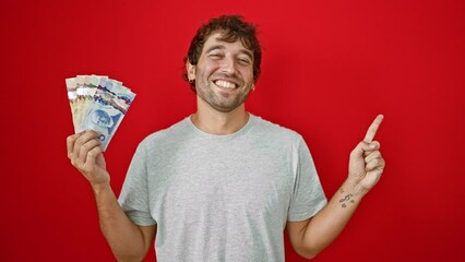 Sticker - Hopeful young man joyfully holding canadian dollar banknotes, confidently pointing to the side with a friendly smile, isolated on a red background.