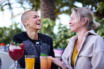 Mature cheerful Caucasian sisters meeting sitting on terrace of restaurant drinking cocktail talking and smiling. Two middle-aged adult friends together enjoying having fun evening at outdoor bar. 