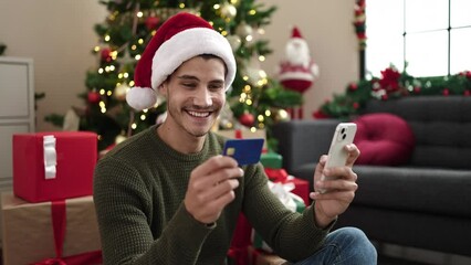 Wall Mural - Young hispanic man using smartphone and credit card sitting on floor by christmas tree at home