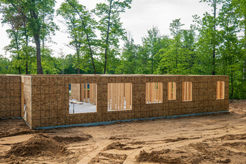 Poster - New home building site in early construction stage, with plywood on exterior walls with window openings and wood framing studs inside, on a wooded lot.