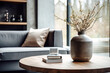 Close up of ceramic vase with blossom twigs on round wooden coffee table against grey sofa and window. Minimalist home interior design of modern living room.