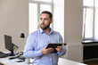 © fizkes - Serious handsome millennial business owner man using tablet computer, standing at office workplace, looking away with concern, thinking over company problems, making decision