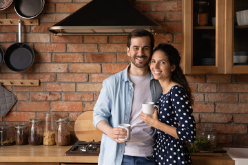 Wall Mural - Family portrait happy young couple drinking coffee or tea in morning, standing in cozy kitchen at home, smiling wife and husband hugging, looking at camera, holding mugs, starting new day together