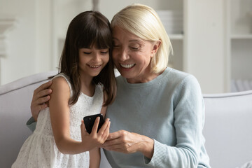 Wall Mural - Happy grandmother and little granddaughter using phone together, having fun at home, sitting on cozy sofa, smiling positive senior granny hugging cute girl grandchild, playing game, browsing apps