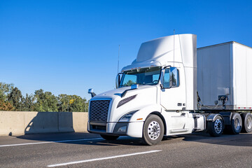 Powerful day cab white big rig semi truck with built-in roof spoiler transporting cargo in dry van semi trailer running on the road at sunny day