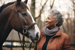 A senior woman standing close to a horse outdoors in nature, holding it