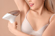 Beautiful young woman epilating her armpits with modern photoepilator on brown background