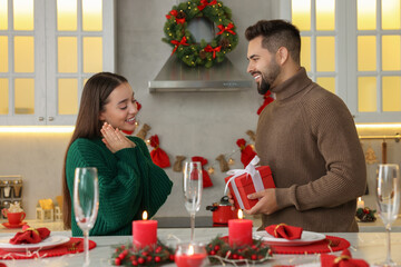 Wall Mural - Happy young man surprising his girlfriend with Christmas gift at table in kitchen