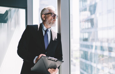 Sticker - Thoughtful senior businessman with clipboard standing near glass wall in office