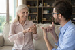 © fizkes - Grown up son communicates with mature retired mother using gestures. Mature woman with hearing loss, physical disability interact with young adult son showing each other symbols sit on sofa at home