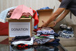 Asian woman putting used clothes into a cardboard box with other used clothes for donation, social assistance, support for victims of war, natural disasters, helping people, poverty and charity.