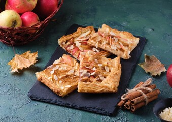 Wall Mural - Sliced open apple pie on puff pastry with apple slices, sprinkled with almonds, on a black slate board on a green concrete background. Recipes apple pie