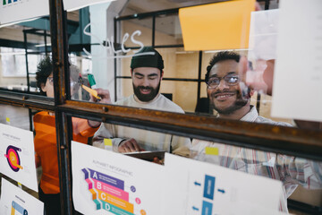 Wall Mural - Smiling colleagues working on plan of project in workspace with stickers on glass wall