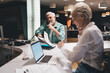 © BullRun - Joyful interaction between a senior Caucasian woman and a man in a casual office setting. The woman laughing using laptop, man is speaking on phone holding documents, collaborative work environment.