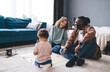 © BullRun - Happy diverse family with kid sitting on floor near sofa at home