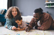 © BullRun - Happy diverse family with little kid lying on floor with map in living room