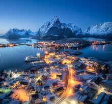 Aerial View Of Snowy Village, Islands, Rorbu, City Lights, Blue Sea, Rocks And Mountain At Night In Winter. Beautiful Landscape With Town, Street Illumination. Top View. Reine, Lofoten Islands, Norway