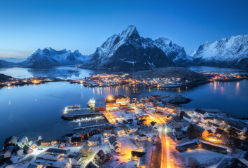 Wall Mural - aerial view of snowy village, islands, rorbu, city lights, blue sea, rocks and mountain at night in 