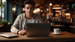 A young man working on a laptop at a cafe table, facing the camera,