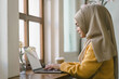 © kenchiro168 - Millennial Asian Muslim freelancer businesswoman working remotely online using a laptop at a cafe.