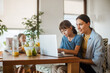 © Halfpoint - Mother working on laptop at kitchen table while children have breakfast. Remote work and home office for mothers with children.
