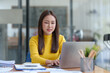 © amnaj - Happy young Asian woman smiling while working with laptop in office.