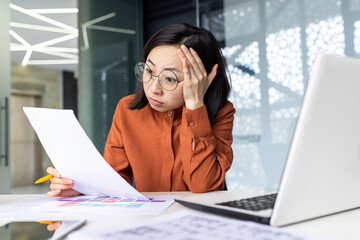 Tired young chinese woman wearing glasses sitting at desk in office with pile of papers in hands, holding head from tiredness and headache, overworked concept.