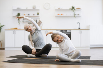 Wall Mural - Slim muslim lady on yoga mat stretching arms while training with small female kid. Young parent and daughter in sportswear spend time happily during workout.