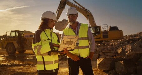 Poster - Caucasian Male Engineer And Hispanic Female Building Architect Walking On Construction Site With Laptop Computer And Talking About New Real Estate Project. Heavy Machinery Working. Golden Hour