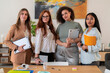 © Studio Marmellata - Group of cheerful young smiling women colleagues in smart elegant clothes standing near wooden table and looking at camera while holding laptops and notebook at office