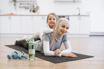 Wall Mural - Adorable girl in sport outfit lying on mothers back with positive energy during exercises. Loving muslim female in hijab relaxing with cute daughter after fitness routine at home.