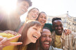 © CarlosBarquero - Happy smiling multi-ethnic group friends having fun together. Multiracial diverse young colleagues embracing posing for selfie photo on street in city. Multicultural friendship generation Z concept.