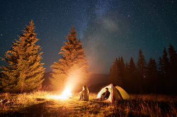 Wall Mural - Night camping in mountains under starry sky. Two people, couple having a rest near burning campfire, enjoying beautiful sky full of stars. Concept of tourism and healthy living.