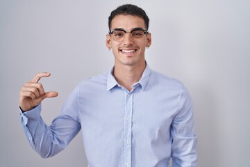 Wall Mural - Handsome hispanic man wearing business clothes and glasses smiling and confident gesturing with hand doing small size sign with fingers looking and the camera. measure concept.