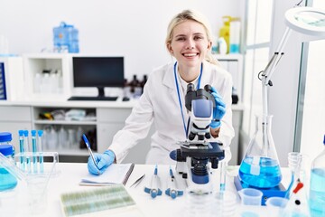 Poster - Young blonde woman scientist using microscope taking notes at laboratory