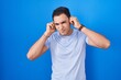© Krakenimages.com - Young hispanic man standing over blue background covering ears with fingers with annoyed expression for the noise of loud music. deaf concept.
