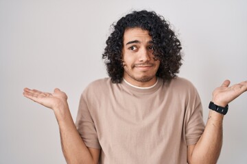 Sticker - Hispanic man with curly hair standing over white background clueless and confused expression with arms and hands raised. doubt concept.