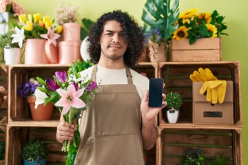 Wall Mural - Hispanic man with curly hair working at florist shop showing smartphone screen skeptic and nervous, frowning upset because of problem. negative person.