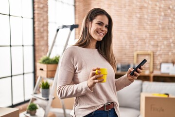 Wall Mural - Young beautiful hispanic woman using smartphone drinking coffee at new home